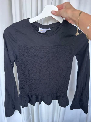 Svart långärmad topp med volang - Säljer en svart långärmad topp med smockad front och volangkant nedtill. Toppen har en rund halsringning och är figurnära i modellen. Perfekt att styla med både jeans och kjol för en trendig look.