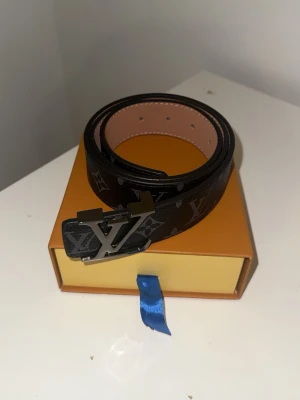Svart Louis Vuitton monogram skärp - Snyggt svart skärp från Louis Vuitton med klassiskt monogrammönster och en stor LV-logga i metall som spänne. Skärpet har en elegant design och är tillverkat i skinn med detaljer i silverfärg. Perfekt accessoar för att lyfta din outfit.