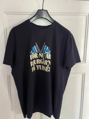 Svart Djurgården t-shirt med tryck - Cool svart t-shirt med tryck fram och bak. Framsidan har texten 'För nu är Djurgår'n på turné!' och två blå-randiga flaggor. Baksidan har en illustration av en person i blå-randig tröja med bengal och texten 'Se bengalernas sken'. Perfekt för Djurgården-supportern.
