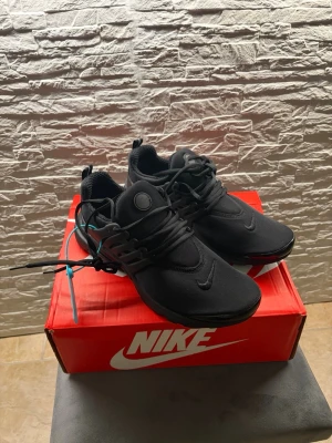Svarta Nike Presto sneakers - Snygga helsvarta Nike Presto sneakers med minimalistisk design och diskret swoosh på tå och plös. Skorna har en flexibel mesh-överdel, plastdetaljer vid snörningen och en bekväm, låg siluett. Perfekta för dig som gillar stilrena och sportiga sneakers.