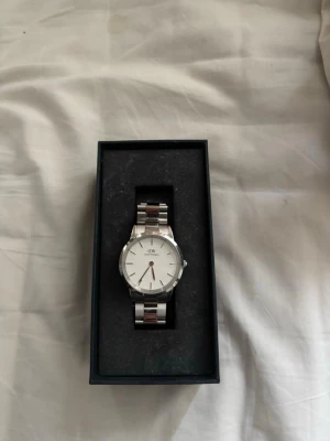 Silverfärgad klocka från Daniel Wellington - Stilren klocka från Daniel Wellington med silverfärgat metallarmband och vit urtavla. Klassisk design med minimalistiska markeringar och DW-logga på urtavlan. Perfekt accessoar för dig som gillar enkel och tidlös stil. Klockan är välomhändetagen, inte använd på länge. Inga repor eller andra skador.
