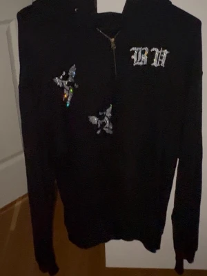 Bvaldi bling hoodie  - Svart hoodie med dragkedja, huva och detaljerade fjärilar framtill. På bröstet finns texten 'GH' i glittrande stil och på ryggen står det 'GUARDIANS' i samma stil. Perfekt för dig som gillar streetwear och unika detaljer. Sjukt snygg och storlek S, köptes för 1049kr