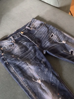 Blå jeans distressed  - Säljer ett par blå jeans med slitningar, hål (distressing) och färgstänk. Jeansen har klassisk femficksmodell, perfekta för dig som gillar en trendig och avslappnad stil. 