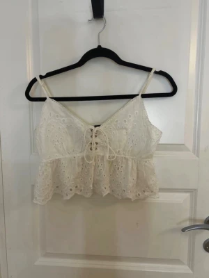 Vit broderad linnetopp med snörning - Söt vit linnetopp med broderade detaljer och snörning framtill. Toppen har tunna axelband och volangkant nedtill. Perfekt för varma dagar och ger en romantisk vibe till din outfit.