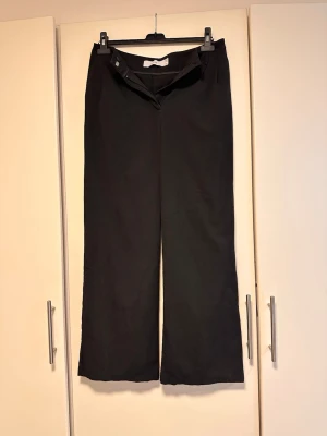 Svarta vida kostymbyxor - Snygga svarta kostymbyxor med hög midja och vida ben. Klassisk design med dragkedja och knapp framtill. Perfekta för en stilren och trendig look. Materialet ser ut att vara mjukt och följsamt, vilket ger en bekväm känsla hela dagen.