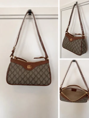 Helt ny Axelremsväska brun  - Snygg axelremsväska i beige och brunt canvasmönster med bruna läderdetaljer. Väskan har guldfärgad logga framtill, dragkedja upptill och justerbar axelrem. Perfekt för dig som gillar klassisk och trendig design.