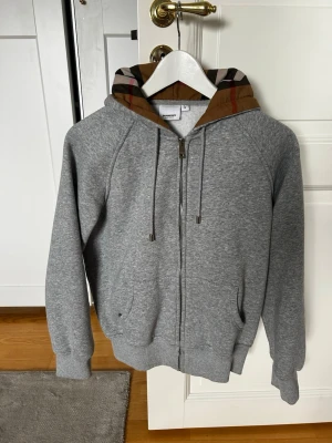 Grå burberry hoodie med mönstrad huva - Snygg grå zip-hoodie med klassisk rutmönstrad huva i brunt, svart, vitt och rött. Hoodien har dragsko i huvan, två fickor fram och ribbade muddar vid ärmar och nederkant. Perfekt för en avslappnad och trendig look. Storlek L men funkar för M. 