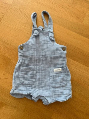 Ljusblå hängselshorts i bomull baby  - Säljer ett par ljusblå hängselshorts i mjuk, krinklad bomull med två stora fickor framtill och knappar på axelbanden. Perfekt för en avslappnad och söt look. Passar till varma dagar och är superbekväma.