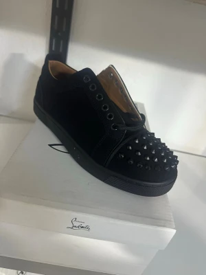 Svarta sneakers med nitar - Säljer ett par svarta sneakers från Christian Louboutin med coola svarta nitar på tån och den ikoniska röda sulan. Skorna har en låg profil och är gjorda i mocka och läder för en lyxig känsla. Perfekta för dig som vill sticka ut med din stil.