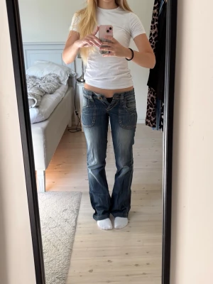 Blå bootcut jeans med stora fickor - Säljer ett par as snygga blå bootcut jeans med stora framfickor och ljusa slitningar som tyvärr är för små för mig. Jeansen har låg midja och kontrastsömmar som ger en snygg retrovibe. Perfekta för dig som gillar en avslappnad men trendig stil. Den enda storleken som står på jeansen är 8/32. Jeansen är i mycket bra skick men har några slitningar längst ner.
