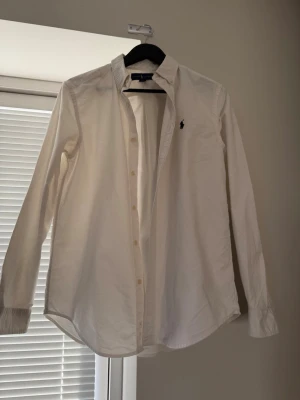 Ralph Lauren skjorta - Klassisk vit skjorta med lång ärm och knappar framtill. Skjortan har en enkel och stilren design med liten broderad logga på bröstet. Perfekt till många olika looks och tillfällen.