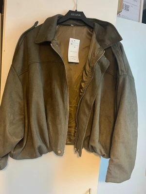 Olivgrön oversized jacka från Indiska - Snygg och trendig olivgrön jacka från Indiska i storlek M. Jackan är oversized med breda ärmar och har en mjuk, mockaliknande yta. Dragkedja framtill och klassisk krage. Perfekt för höst och vår! Lappen sitter kvar, aldrig använd.