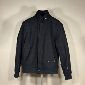 G-star RAW Svart jacka med hög krage och knappar - Snygg svart jacka med hög krage och knappdetaljer. Jackan har två snedställda fickor framtill och muddar vid ärmslut och nederkant. Stängs med dragkedja och knappar. Perfekt för dig som vill ha en stilren och enkel look.0190
