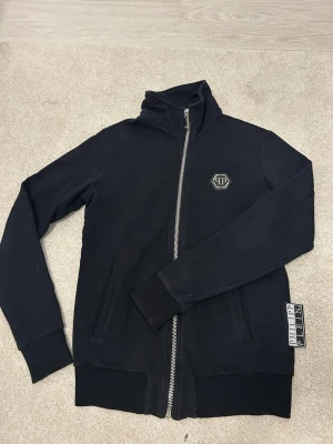  Philipp Plein kofta - Snygg svart hoodie med dragkedja från Philipp Plein. Framsidan har en diskret logga på bröstet och baksidan pryds av en stor, detaljerad tiger i färg. Hoodien har ribbade muddar, hög krage och två fickor framtill. Perfekt för dig som gillar statement-plagg.