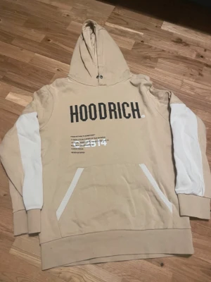 Beige HOODRICH hoodie med vita detaljer - Snygg beige hoodie från HOODRICH med vita paneler på ärmar och nedre delen av ryggen. Stor HOODRICH-logga framtill och världskarta med text på ryggen. Praktisk magficka och huva med dragsko. Perfekt för en streetwear-look.