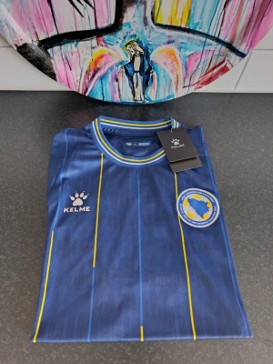 Blå Bosnien & Hercegovina matchtröja (Kelme) – Helt ny med lappar! - ​🇧🇦 Säkra din look inför matchen på Strawberry Arena! 🇧🇦 ​Säljer denna helt nya Bosnien & Hercegovina landslagströja från Kelme. Tröjan är helt oanvänd och alla lappar sitter kvar. ​Snygg design i kungsblått med vertikala gula ränder, fina detaljer på krage och ärmslut samt tryck/märke på bröstet. På baksidan av kragen står texten "BOSNA I HERCEGOVINA". ​🔥 Perfekt att köpa nu innan matchen Sverige möter Bosnien på Strawberry! Visa ditt stöd på plats med en helt ny, fräsch tröja.