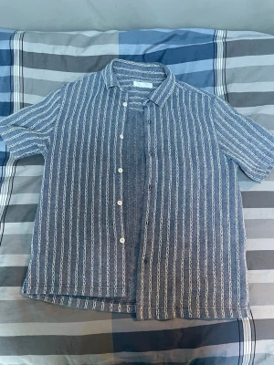 Blåvit randig skjorta i linne - Säljer en blå skjorta med vita vertikala ränder. Skjortan har korta ärmar, klassisk krage och knäppning framtill med vita knappar. Tillverkad i ett luftigt linnematerial som passar perfekt till varma dagar. Enkel och stilren design.