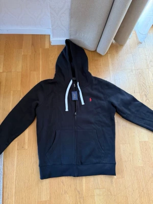 Svart hoodie från Polo Ralph Lauren - Svart hoodie med dragkedja från Polo Ralph Lauren. Klassisk design med vit dragsko i huvan och röd broderad logga på bröstet. Mjuk bomullskvalitet och känguruficka framtill. Perfekt för en avslappnad och stilren look.