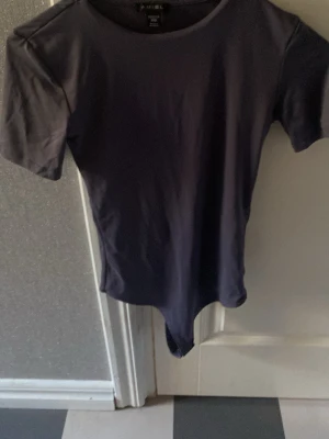 Mörkgrå kortärmad body - En enkel och stilren mörkgrå body med korta ärmar och rund halsringning. Tillverkad i mjukt stretchigt material som sitter skönt mot kroppen. Perfekt att matcha med jeans eller kjol för en clean look.