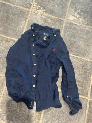 Marinblå linneskjorta från Polo Ralph Lauren - Säljer en marinblå skjorta från Polo Ralph Lauren i svalt linne. Skjortan har klassisk krage, vita knappar och den ikoniska röda logotypen broderad på bröstet. Perfekt för en stilren och avslappnad look.