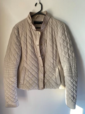 Beige quiltad jacka med ståkrage från Zara - Snygg beige quiltad jacka med ståkrage och guldfärgade knappar. Jackan har långärmad design, fickor med dragkedja och ett rutigt quiltat mönster. Perfekt för kyligare dagar och ger en stilren look.