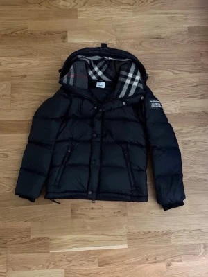 Svart dunjacka/väst med avtagbara ärmar och huva - Snygg svart dunjacka från Burberry London England med avtagbara ärmar och huva. Jackan kan användas både som jacka och väst. Klassisk Burberry-rutig insida på huvan. Två fickor med dragkedja framtill. Perfekt för höst och vinter! Storlek M.