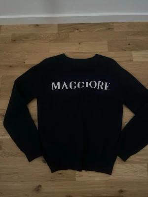 Svart stickad tröja med text - Svart stickad tröja med vit text 'MAGGIORE' över ryggen. Tröjan har rund halsringning och långa ärmar. Enkel och stilren design som passar till mycket. Materialet ser ut att vara mjukt och bekvämt.