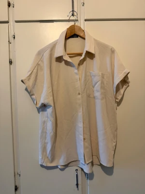 Beige kortärmad skjorta med ficka - Säljer en beige skjorta i oversized modell med korta ärmar och en bröstficka. Skjortan har klassisk krage och knäppning framtill. Perfekt för en avslappnad och trendig look. Ej kommit till användning helt ny. Från SHEIN 