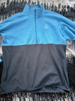 Blå och mörkgrå fleece från Haglöfs - Mysig fleece från Haglöfs i tvåfärgad design med ljusblå upptill och mörkgrå nedtill. Tröjan har halv dragkedja framtill och hög krage. Perfekt för lager-på-lager och outdoor-häng.