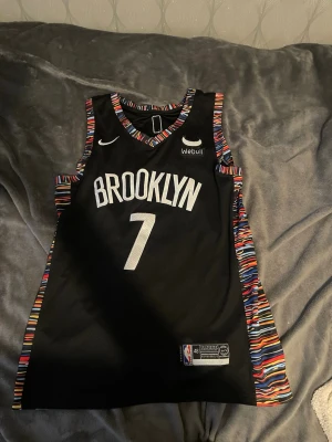 Brooklyn Nets basketlinne #7 Durant - Snyggt svart basketlinne från Brooklyn Nets med nummer 7 och Durant på ryggen. Linnet har färgglada ränder längs kanterna och är tillverkat i ett lätt och ventilerande material. Perfekt för basketträning eller att bära som streetwear.