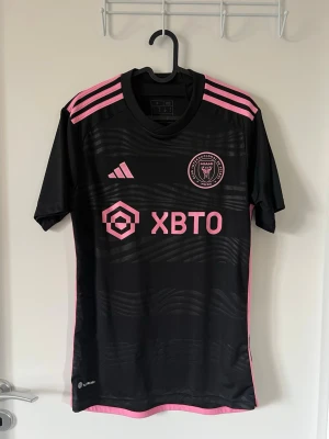 Inter Miami Messi matchtröja svart/rosa storlek S adidas - Snygg svart Inter Miami matchtröja med rosa detaljer och Messi 10 på ryggen. Tröjan har klubbmärke och sponsortryck framtill, samt rosa ränder på axlarna. Tillverkad i ett lätt och ventilerande material, perfekt för fotboll eller träning. Den är oanvänd och köpt på adidas hemsida vid släppet av kollektionen. Det står inte ”man eller kvinna” på men modellen är slim.