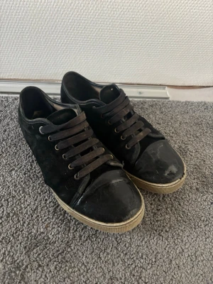 Lanvins - Säljer ett par svarta sneakers med mockadetaljer på sidorna och tåparti i blankt material. Skorna har svarta skosnören och en beige sula som ger en snygg kontrast. Klassisk låg modell som passar till det mesta.