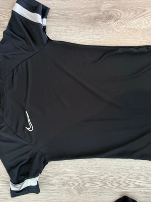 Svart Nike sporttröja med vita detaljer - Säljer en svart sporttröja med korta ärmar och vita detaljer på ärmsluten. Tröjan har en sportig passform och är tillverkad i ett lätt, snabbtorkande material som passar perfekt till träning eller match.