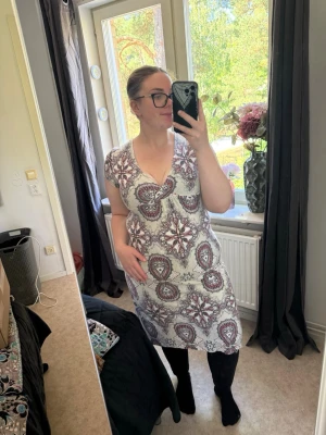 Vit mönstrad klänning med v-ringning - Klänning i vitt med lila och grått paisleymönster. Klänningen har v-ringad hals, korta ärmar och en avslappnad passform. Perfekt för dig som gillar boho-vibbar och vill ha något bekvämt men ändå snyggt. Från märket Odd Molly i storlek 2 som enligt deras storlekguide motsvarar en storlek M. Jag har vanligtvis en 4/XL. Men ör Myclet trång i armhåla och magen, Passar en M/L. Mycket fint skick, jag hittar inga synliga anmärkningar eller defekter.  