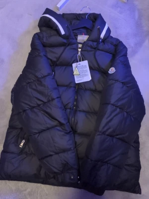 Svart pufferjacka med huva - Snygg svart pufferjacka med huva och dragkedja framtill. Jackan har vita detaljer på huvan och två fickor med dragkedja. Perfekt för kalla dagar och har en klassisk quiltad design. Moncler-logga på ärmen ger en exklusiv touch. Dragkedja på huvan, när den är öppen står de "Moncler"