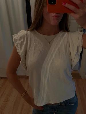 Vit topp med volangärmar - Säljer en söt vit topp med volangärmar och rund hals. Toppen har en lös passform och är tillverkad i ett mjukt bomullstyg. Perfekt att styla med jeans eller kjol för en fräsch look.