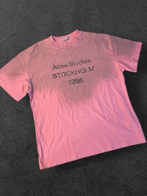 Rosa Acne Studios t-shirt med tryck | S/M - Säljer en rosa t-shirt från Acne Studios med svart texttryck 'Acne Studios STOCKHOLM 1996' på bröstet. Klassisk passform med rund hals och korta ärmar. Perfekt för en trendig och avslappnad stil.