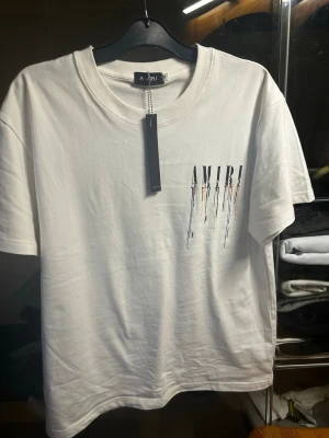 Vit AMIRI t-shirt  - Säljer en vit t-shirt från AMIRI med tryck på bröstet där loggan har en cool drip-effekt. Klassisk rund hals och korta ärmar. Perfekt för dig som gillar streetwear och vill ha något stilrent men ändå unikt.