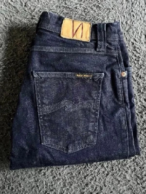 Nudie jeans  - Snygga mörkblå skinny jeans från Nudie med klassisk femficksdesign och läderpatch bak i midjan. Jeansen är i stretchigt denim och har en smal passform som sitter tight längs benen. Perfekta för dig som gillar en stilren och enkel look. Längd 112cm. Ifall det inte finns någon bild i annonsen där jag bär plaggen jag säljer så är det för att jag ej kan ha storleken heller passformen, så kolla måtten för storlek och googla på modellnamnet för passform 😉(n4)