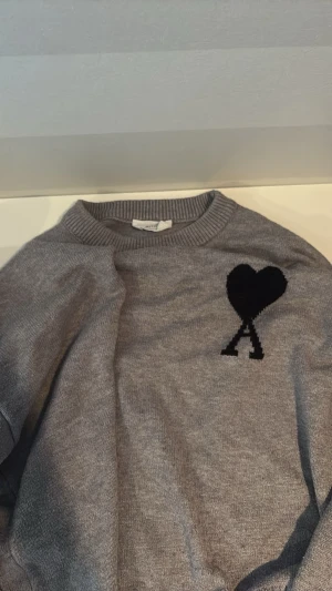 Ami Paris tröja - Snygg grå stickad tröja med svart hjärta och bokstaven A på bröstet. Tröjan har rund halsringning och ribbade muddar vid ärmslut och nederkant. Perfekt för dig som gillar enkel men trendig stil.