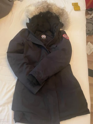 Svart dunjacka från Canada Goose - Säljer en svart dunjacka från Canada Goose med klassisk patch på ärmen och stor huva med fluffig pälsimitation. Jackan har hög krage, dragkedja och knappar framtill samt långa ärmar. Perfekt för kalla vinterdagar.
