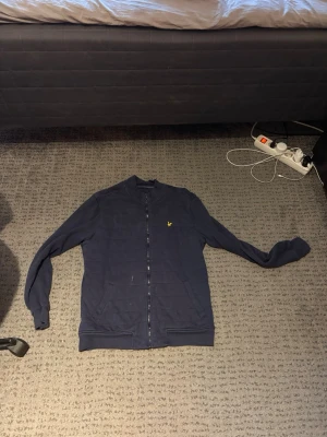 Mörkblå ziptröja från Lyle & Scott - Snygg mörkblå ziptröja från Lyle & Scott med gul broderad logga på bröstet. Tröjan har ribbade muddar, ståkrage och dragkedja hela vägen. Perfekt till chill eller streetwear-stil.