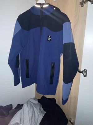 Blå och svart Nike Air Max vindjacka - Snygg vindjacka från Nike Air Max i blått och svart med dragkedja framtill och två fickor med svarta dragkedjor. Jackan har huva och logga på bröstet samt texten 'AIR MAX' på ryggen. Perfekt för en sportig och avslappnad stil.