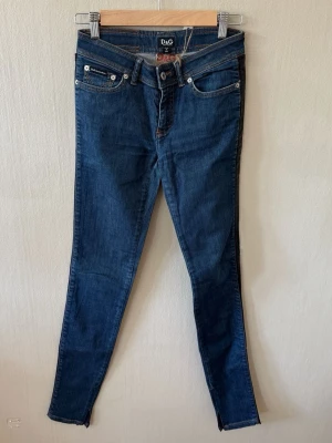Mörkblå skinny jeans från D&G - Snygga mörkblå skinny jeans från D&G med klassiska fem fickor och silverfärgade detaljer. Jeansen har svarta detaljer längs sidorna och smal passform som sitter tight hela vägen ner. Perfekta för dig som gillar en stilren och trendig look. 0098