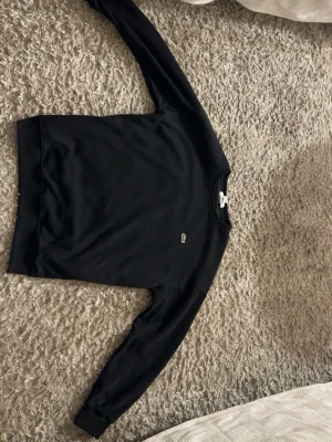 Svart sweatshirt från Lacoste - En stilren svart sweatshirt från Lacoste med rund halsringning och diskret krokodillogga på bröstet. Tröjan har långa ärmar och ribbade muddar vid ärmslut och nederkant. Perfekt för en avslappnad och trendig look.