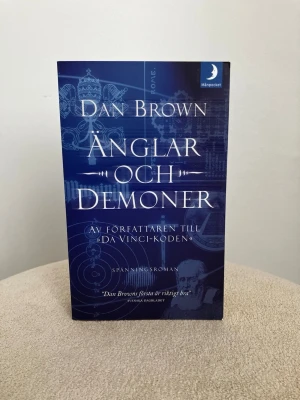 Änglar och Demoner av Dan Brown - Spännande roman av Dan Brown i pocketformat. Omslaget är mörkblått med vita och ljusblå detaljer samt grafiska mönster och text. Boken handlar om konspirationer, mysterier och religiösa symboler i Rom. Perfekt för dig som gillar thrillers och äventyr.