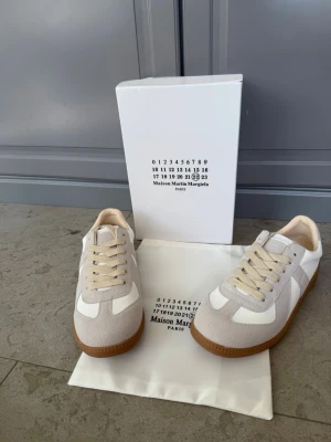 Maison Margiela vita sneakers med beige detaljer - Maison Margiela sneakers i vitt läder och beige mocka med klassisk gummisula. Lågt skaft och stilren design med ton-i-ton snörning. Perfekta för dig som gillar minimalistisk och trendig stil.