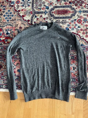 Grå stickad tröja från Hollister - Mysig grå stickad tröja från Hollister med rund halsringning och diskret logga broderad på bröstet. Tröjan har långa ärmar och ribbade muddar vid ärmslut och nederkant. Perfekt för lager-på-lager och enkel att matcha.