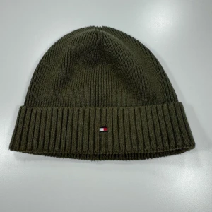 Tommy Hilfiger Ribbad Mössa / Unisex One Size / Olivgrön - Storlek enligt märkning: OS (One Size) Passar: One Size / Unisex  Mått (plagget liggande platt): Bredd: 25 cm Längd: 21 cm  Ribbstickad mössa från Tommy Hilfiger i olivgrönt. Modellen har en bred uppvikt kant och en broderad flagg-logotyp fram. Klassisk unisex-modell med rak, tät ribbstickning.  Mycket gott begagnat skick – inga hål, revor eller fläckar. 0381