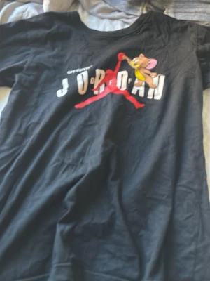 Svart t-shirt med Jerry och Jordan-tryck - Cool svart t-shirt med stort tryck av Jerry från Tom & Jerry som gör en Jordan-dunk. Texten 'JORDAN' syns i vitt och rött över bröstet. T-shirten har rund hals och korta ärmar. Perfekt för dig som gillar streetwear och tecknade motiv.